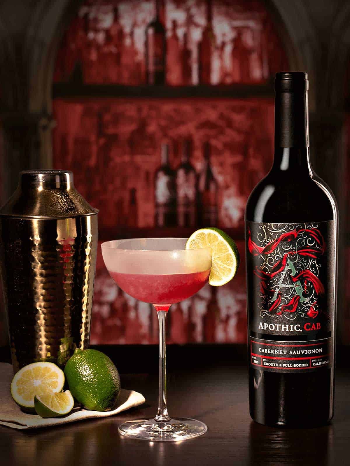 A glass of the bishop cocktail with a shaker, limes and a bottle of Apothic cabernet sauvignon