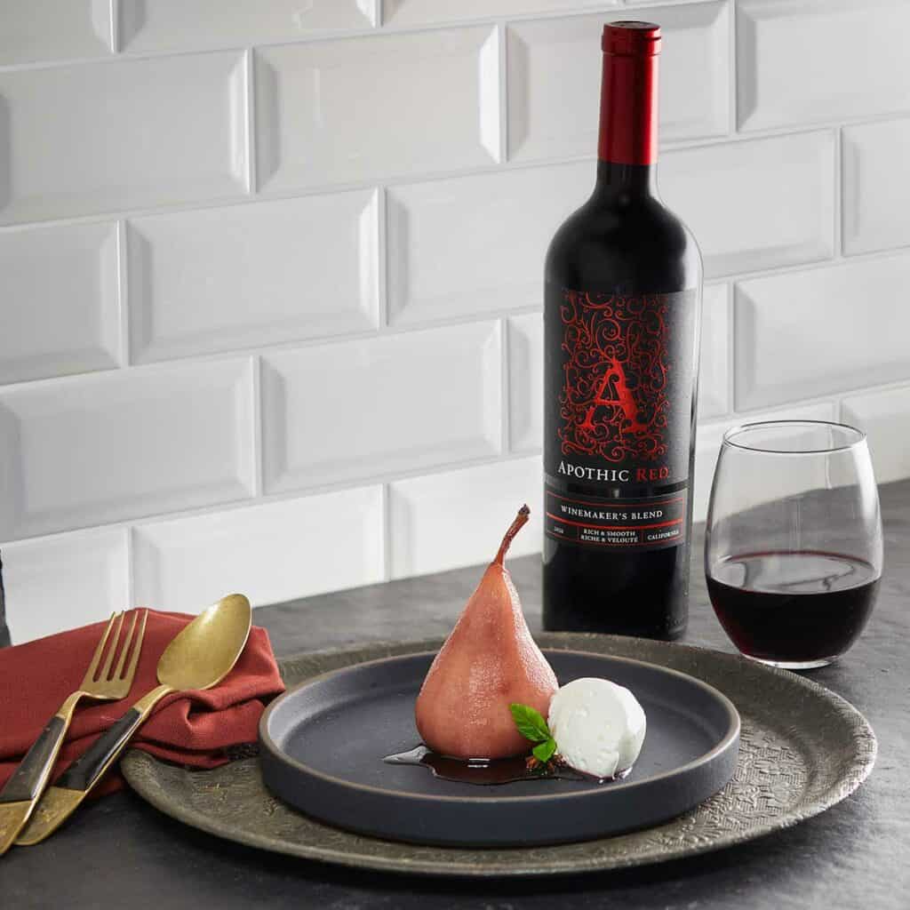 A gray plate with a poached pear and whipped cream in it, in front of a glass and a bottle of Apothic red wine