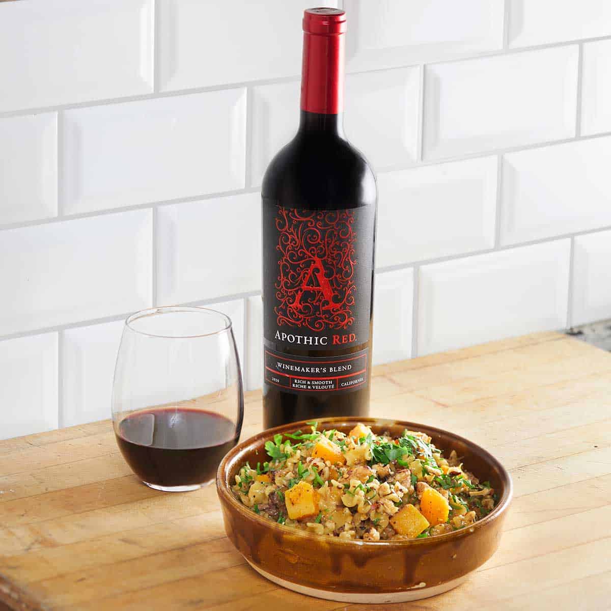 A buttery squash and duck pasta bowl in front of a glass and a bottle of Apothic Red