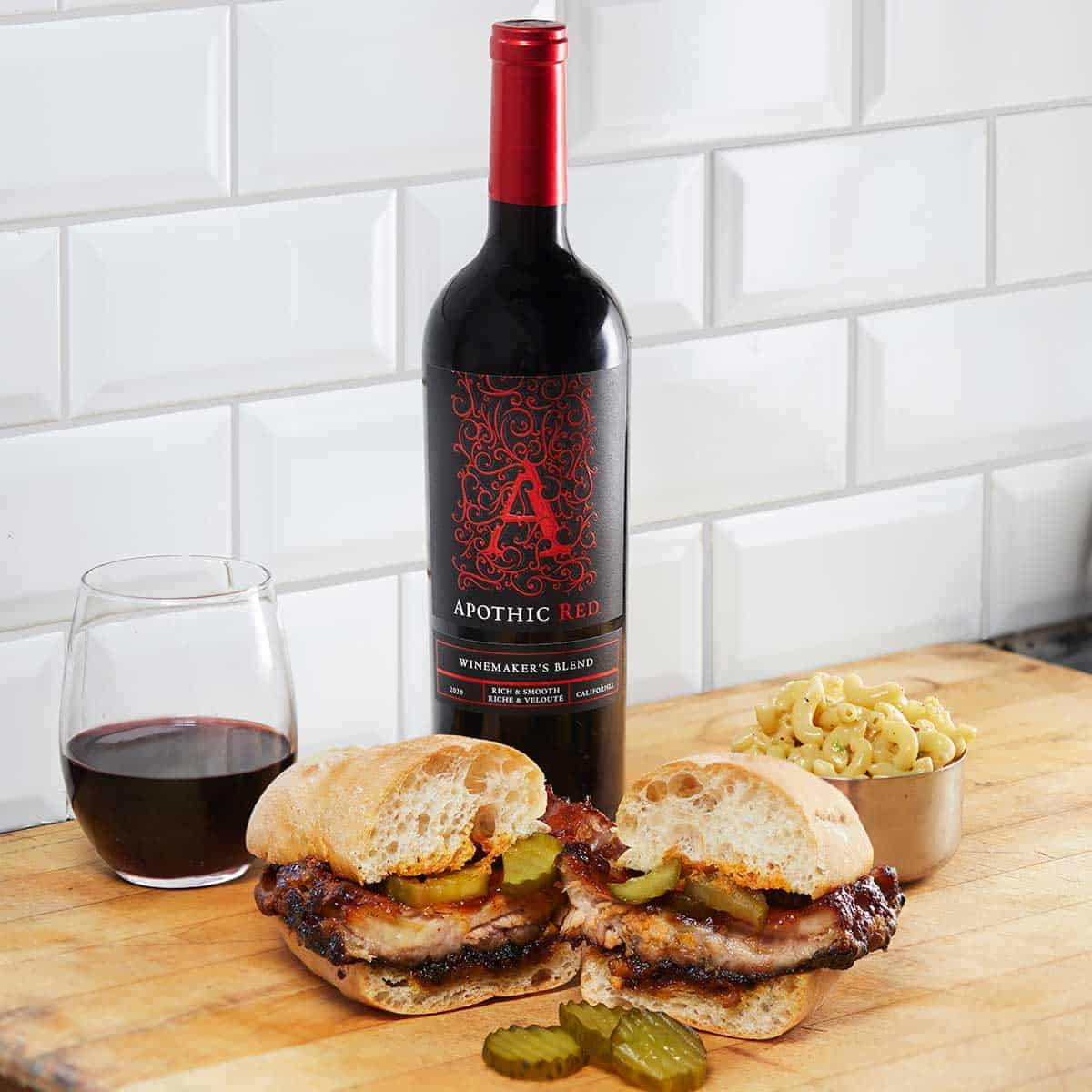 A rib sandwich and pasta salad bowl in front of a glass and a bottle of Apothic Red