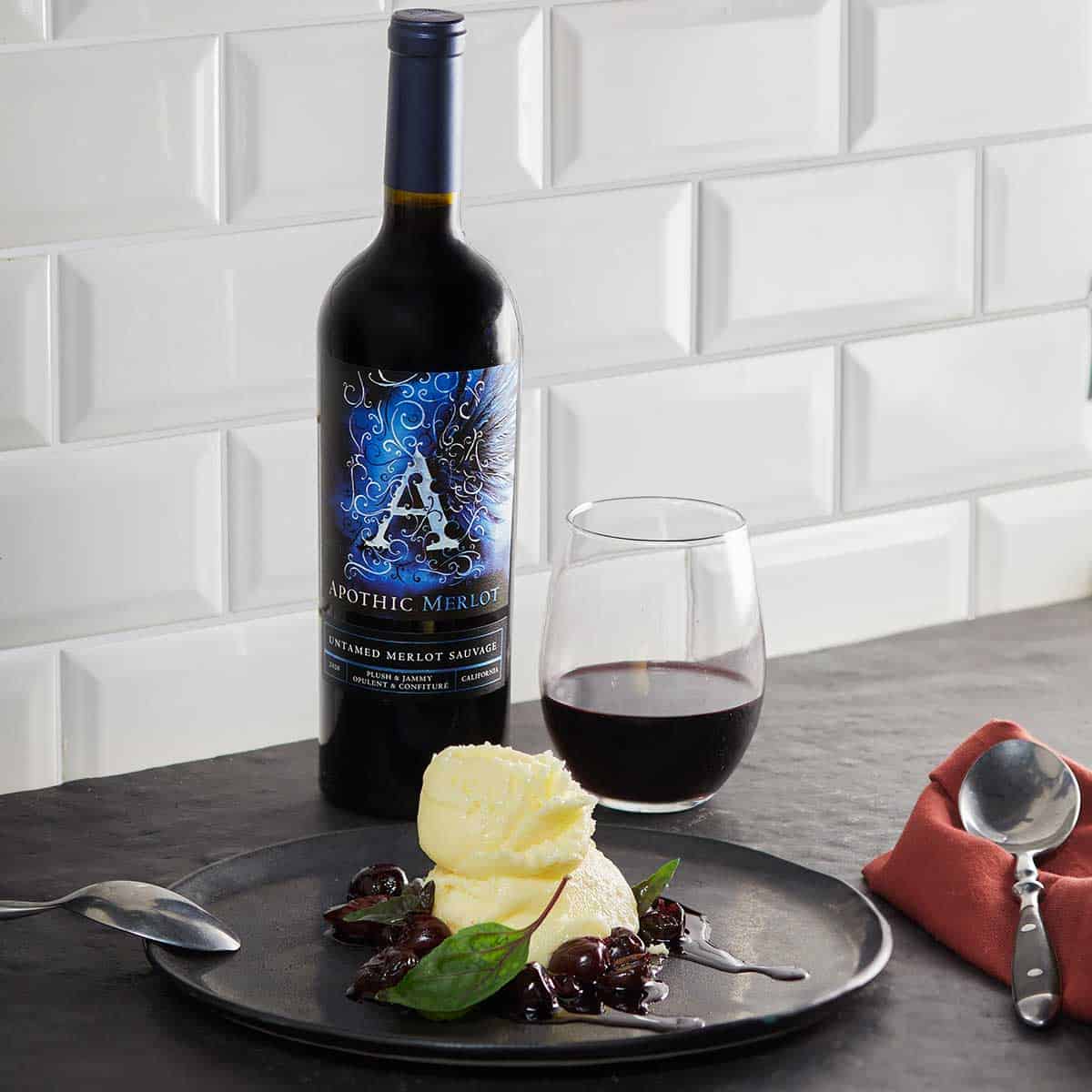 A vanilla ice cream and cherry plate in front of a glass and a bottle of Apothic Merlot