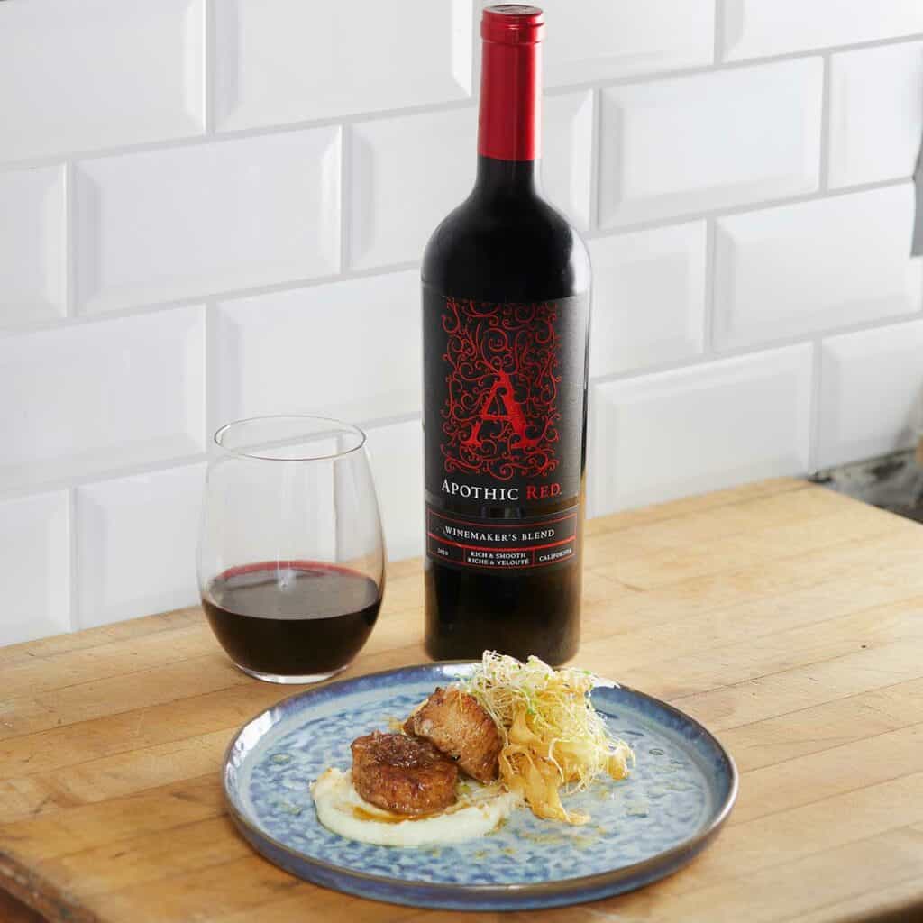 A seared scallops plate in front of a glass and a bottle of Apothic Red