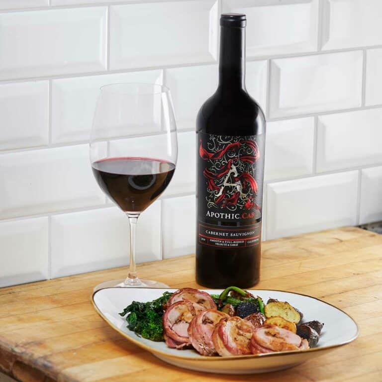 A roast pork loin and vegetables plate in front of a glass and a bottle of Apothic Cab