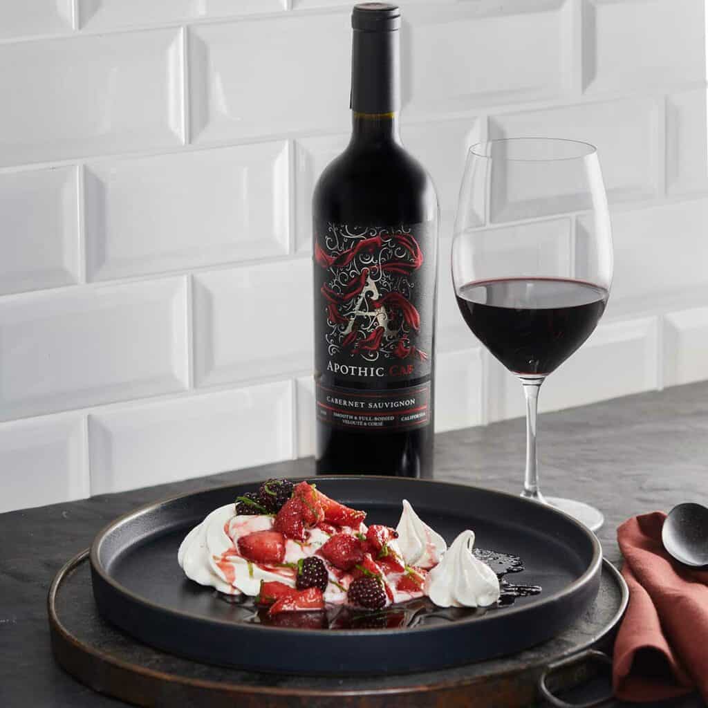 A pavlova with berries and wine jam plate in front of a glass and a bottle of Apothic Cab