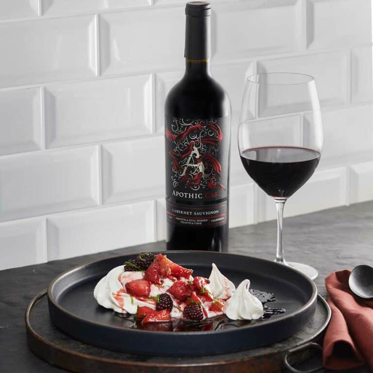 A pavlova with berries and wine jam plate in front of a glass and a bottle of Apothic Cab