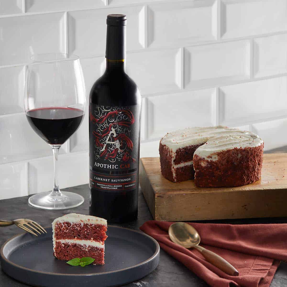 A red velvet cake in a plate in front of a glass and a bottle of Apothic Cab