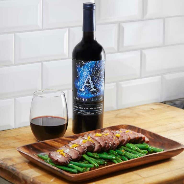 A wooden plate with duck breast on a bed of french beans in front of a glass and a bottle of Apothic Merlot
