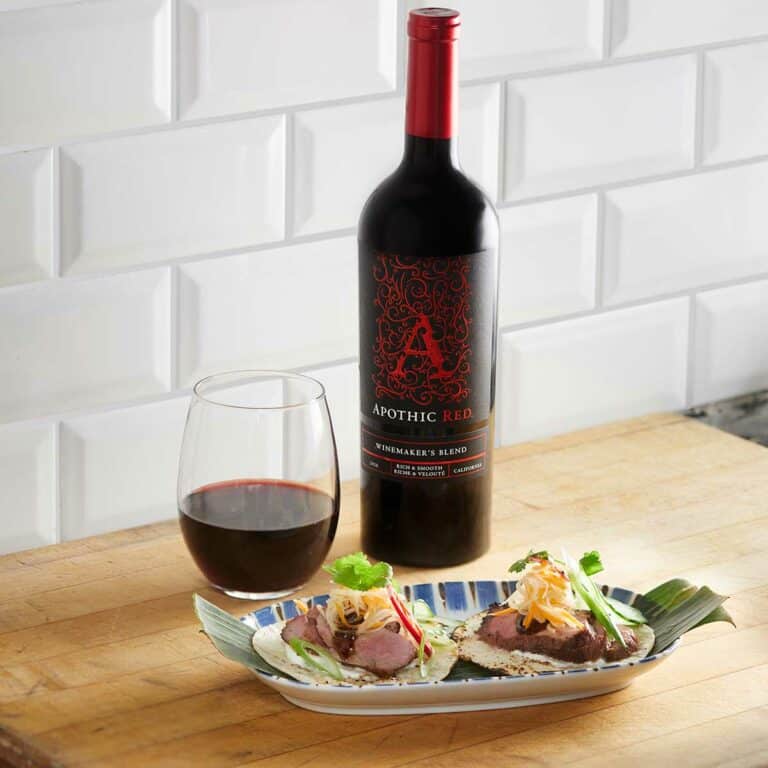 Two duck tacos in a plate in front of a glass and a bottle of Apothic Red