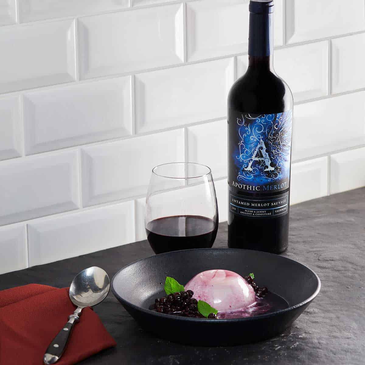 A panacotta bowl with berry wine sauce in front of a glass and a bottle of Apothic Merlot