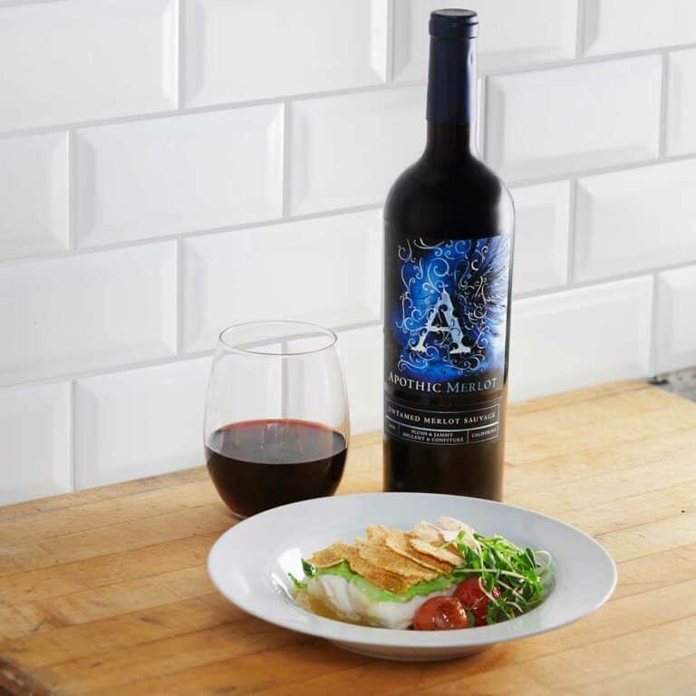 Two seared halibut filets with cornmeal chips and tomatoes in a bowl in front of a glass and a bottle of Apothic Merlot